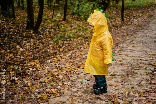 Kid in a yellow raincoat walks through the autumn park on a rainy day. The raincoat has a green crest like a dinosaur