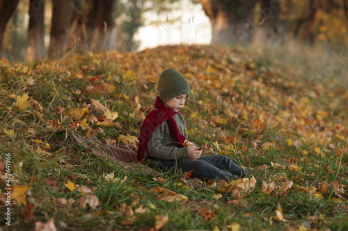 a boy child is sitting on the grass  wearing a green sweater  a red scarf  holding a candy in his hand and looking at it