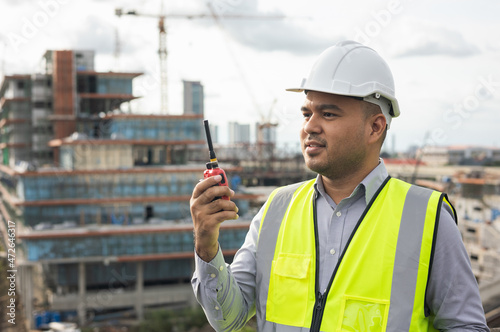 Asian engineer handsome man or architect use radio communication with white safety helmet in city construction site . Standing on rooftop building construction at capital.