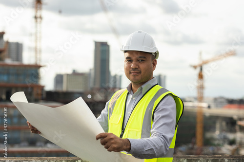Asian engineer handsome man or architect looking blueprint with white safety helmet in city construction site . Standing with paperwork on rooftop building construction at capital.