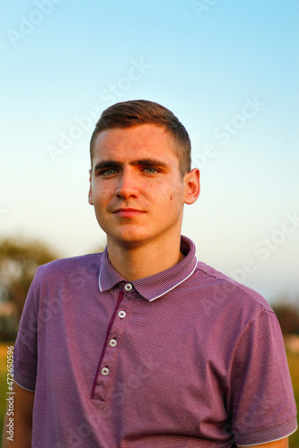 Young people.Portrait of serious young brunette man in purple polo shirt standing outdoor on nature background at summer day. 20s years guy. Millennial generation. Outside