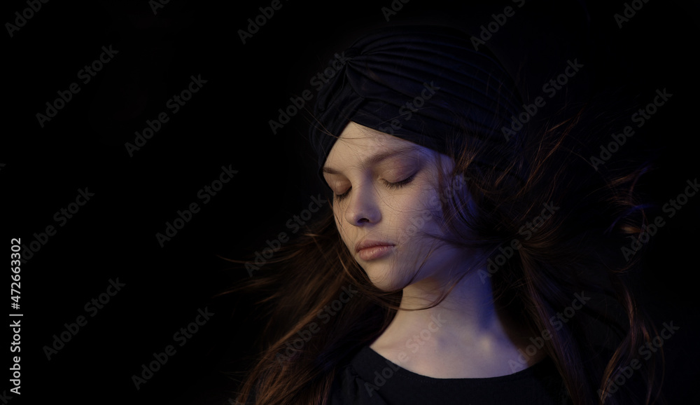 Beautiful little teenager girl with meditative facial expression in multicolor lights. Dark portrait of child with turban and flowing hair shows emotions. Selective focus, low key
