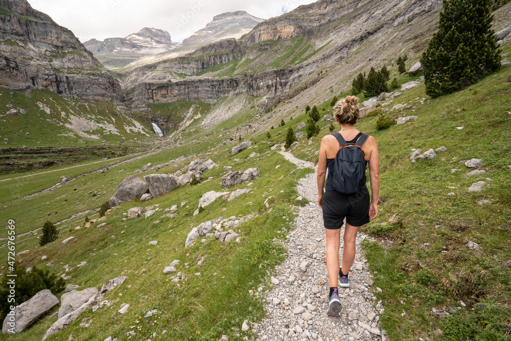 A woman walks through a mountain valley wearing summer clothes. The path ends in a waterfall