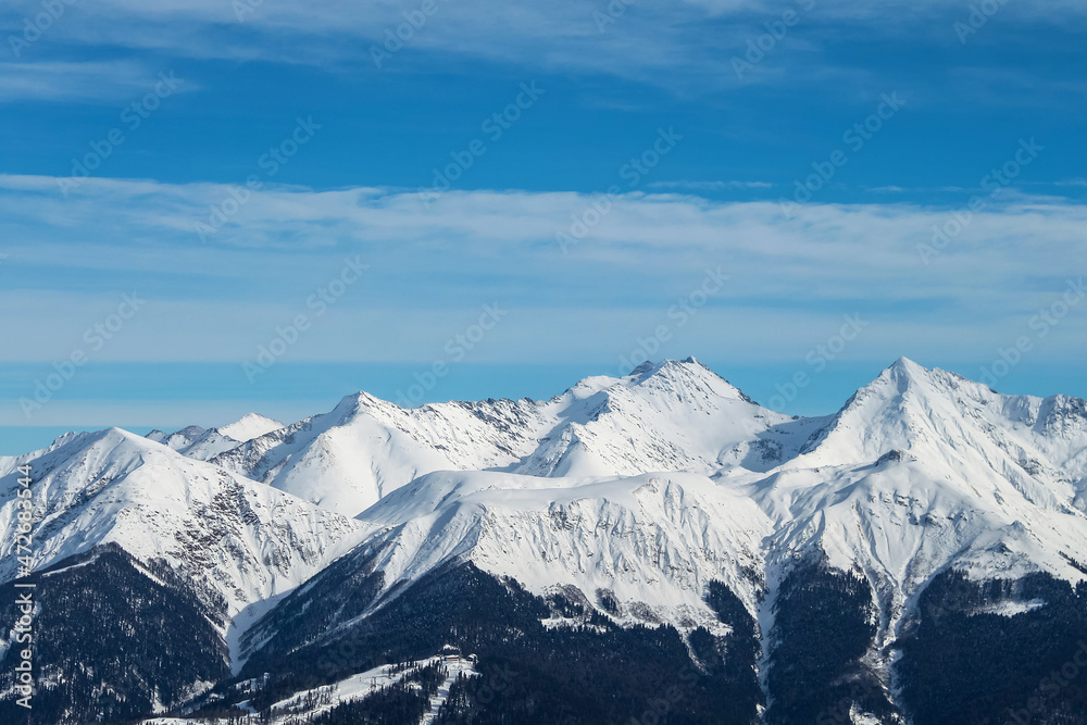 Snowy mountain peaks against a background of blue sky and clouds. The mountains are black from below, while the middle and peaks are completely white. A beautiful winter landscape.