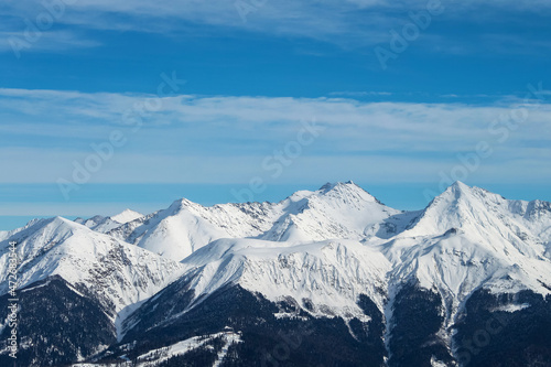 Snowy mountain peaks against a background of blue sky and clouds. The mountains are black from below, while the middle and peaks are completely white. A beautiful winter landscape. © VeNN