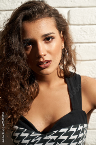 Close up isolated portrait of attractive woman with curly hair posing near white brick wall. Front view of beautiful seductive young female model wearing black and white dress. Concept of beauty.