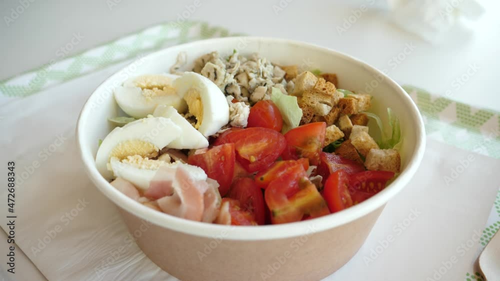 Bowl with fresh Caesar salad of vegetables and beacon meat, eggs and croutons in disposable paper tableware ordered in mall food court Bowl with fresh Caesar salad of vegetables and beacon meat, eggs and croutons in disposable paper tableware ordered in mall food court