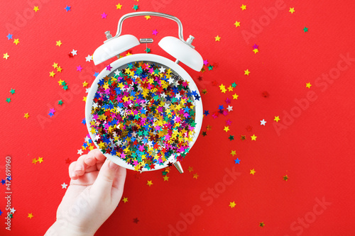 child's hands hold alarm clock with scattered sequins on a red background. christmas, celebration concept