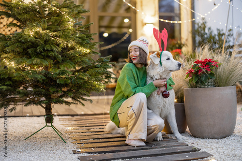 Young woman with her cute white dog at New Year's decorated backyard of country house. Girl having fun with a dog on winter holidays. Dog wearing deer ears