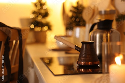 Fototapeta Naklejka Na Ścianę i Meble -  Beautiful copper coffee pot on cooktop in kitchen against blurred Christmas lights, space for text