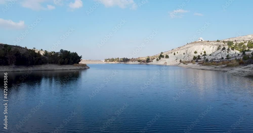 Aerial shot over a water reservoir dam surrounded by small hills and mountains over a blue sky day Aerial shot over a water reservoir dam surrounded by small hills and mountains over a blue sky day