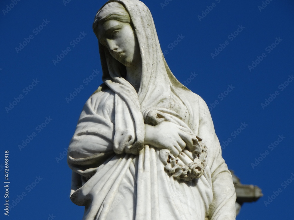 Estátua de marmore que retrata uma mulher de véu com olhar melancólico que em suas mãos está segurando uma bela guirlanda de flores  no meio de um cemitério antigo com lindo céu azul ao fundo.