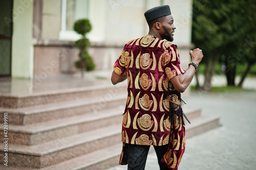 Back of african stylish and handsome man in traditional outfit and black cap standing outdoor.