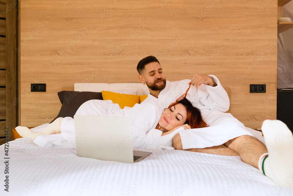 Fototapeta premium A young couple in bathrobes is lying on the bed with a laptop. A man playing with a woman's hair. A woman lies with her head in the lap of a man.