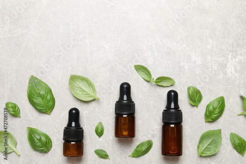 Bottles of essential basil oil and fresh leaves on light grey table, flat lay. Space for text