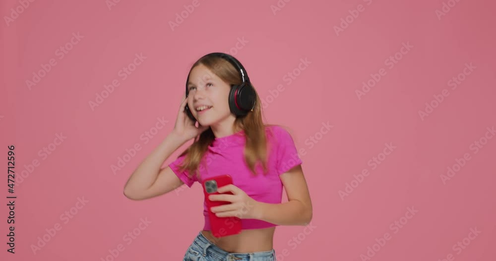 Pretty child girl enjoying music with phone in hand wearing headphones dancing having fun on pink studio background Pretty child girl enjoying music with phone in hand wearing headphones dancing having fun on pink studio background