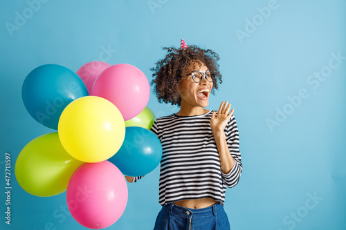 Cheerful multiracial lady in birthday cone hat looking away and laughing while holding multicolored balloons. Isolated on blue background