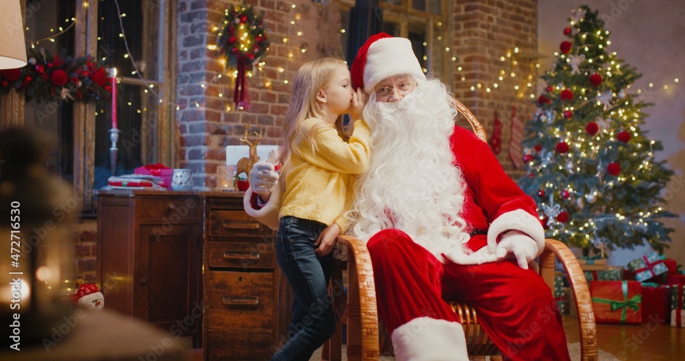 What do you want. Santa Claus sitting at the armchair and listening wishes of little girl standing nearby. New Year and holidays concept What do you want. Santa Claus sitting at the armchair and listening wishes of little girl standing nearby. New Year and holidays concept