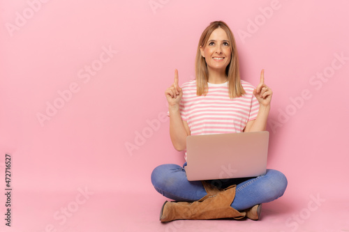 Young smiling woman in striped t-shirt using laptop computer pointing fingers up sitting on floor over pink background.