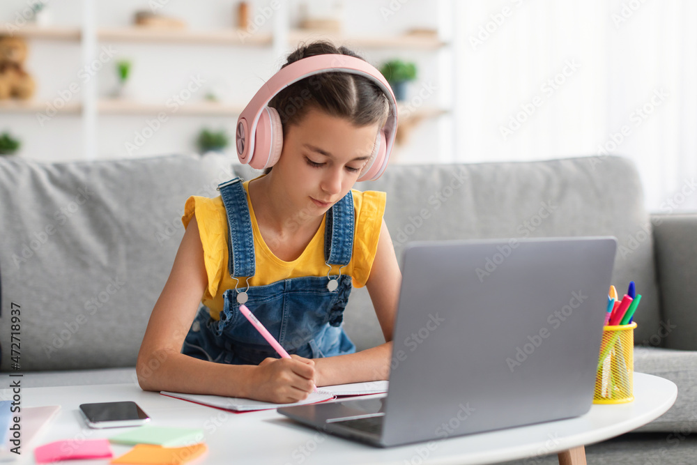 Schoolgirl in headset sitting at desk, writing in textbook Schoolgirl in headset sitting at desk, writing in textbook