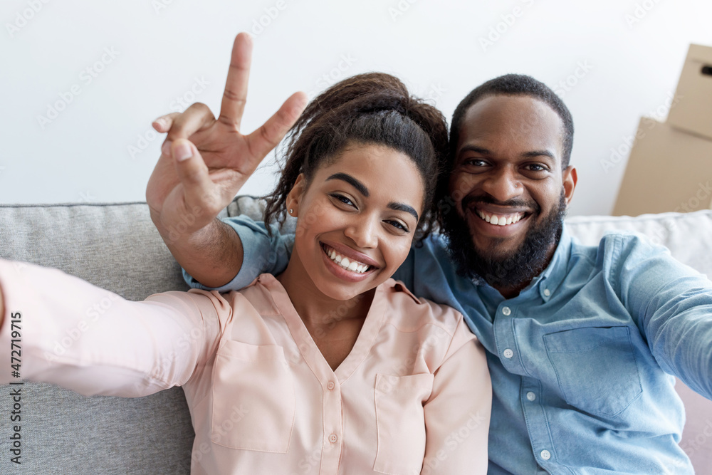 Smiling young black couple rest on couch, man shows peace sign, makes selfie in room Smiling young black couple rest on couch, man shows peace sign, makes selfie in room
