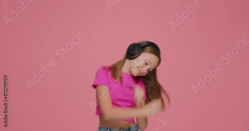 Cute little kid girl listen to music with wireless headphones, dancing, having fun on pink studio background