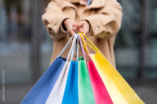 Cropped view of young woman in warm winter coat holding colorful shopping bags outdoors, closeup