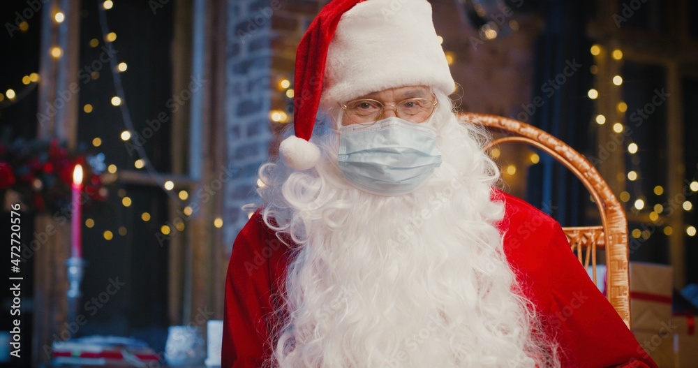 Santa Claus sitting at the armchair and looking at the camera with presents with New Year decorations at the background. Holidays spirit concept Santa Claus sitting at the armchair and looking at the camera with presents with New Year decorations at the background. Holidays spirit concept