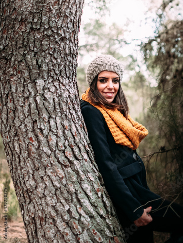 Joven mujer apoyada del tronco de un árbol un día de invierno por el bosque.