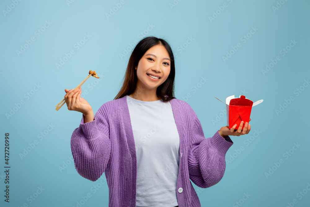 Food delivery, fastfood concept. Asian lady eating noodles with chopsticks, holding paper box over blue background