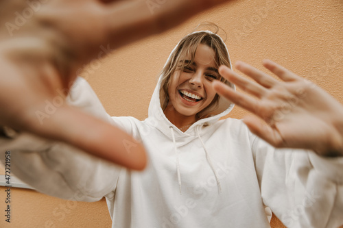 Cheerful young fair-skinned lady hiding from paparazzi hiding behind her hands against background of wall. Blonde with perfect smile wears white sweatshirt. Freedom concept
