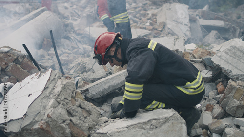 Pan left view of male emergency workers removing concrete rubble in cloud of dust while working on ruins of destroyed building after disaster photo