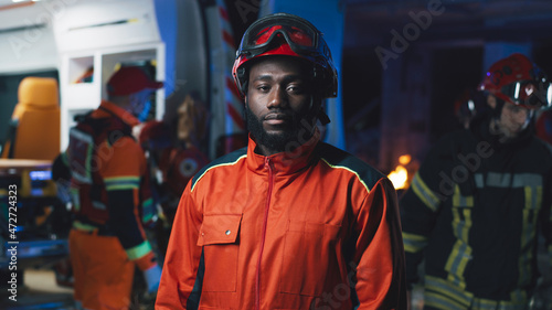 Bearded African American man in hardhat looking at camera while standing near van and colleagues during accident at night