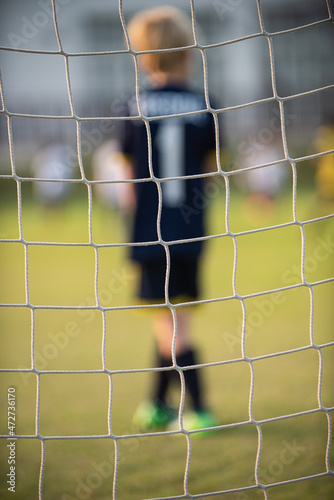 Close up football goal net, seeing a young boy soccer goalkeeper watching his teammate playing. Youth Soccer game on a sunny summer school tournament Day. A football match going on in a background.