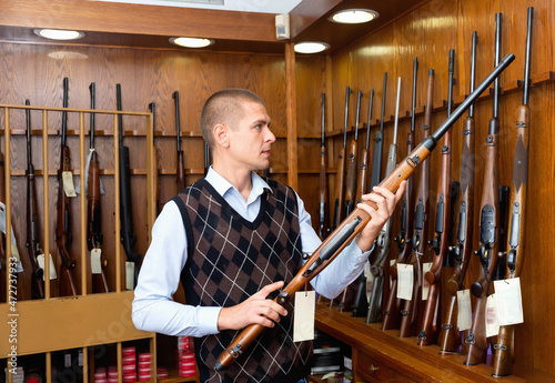 Confident male hunter examining hunting shotgun before buying in gun store.