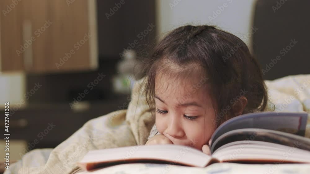 Close up 4K of adorable 5 years old Asian girl who is reading a book on a bed with beautiful yellow light from a lamp. It is bedtime studying for learning, relaxing and developing creativity of child. Close up 4K of adorable 5 years old Asian girl who is reading a book on a bed with beautiful yellow light from a lamp. It is bedtime studying for learning, relaxing and developing creativity of child.