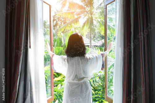 A woman opens a window to welcome the morning sunlight of a new day at her vacation home. Natural tree background. photo