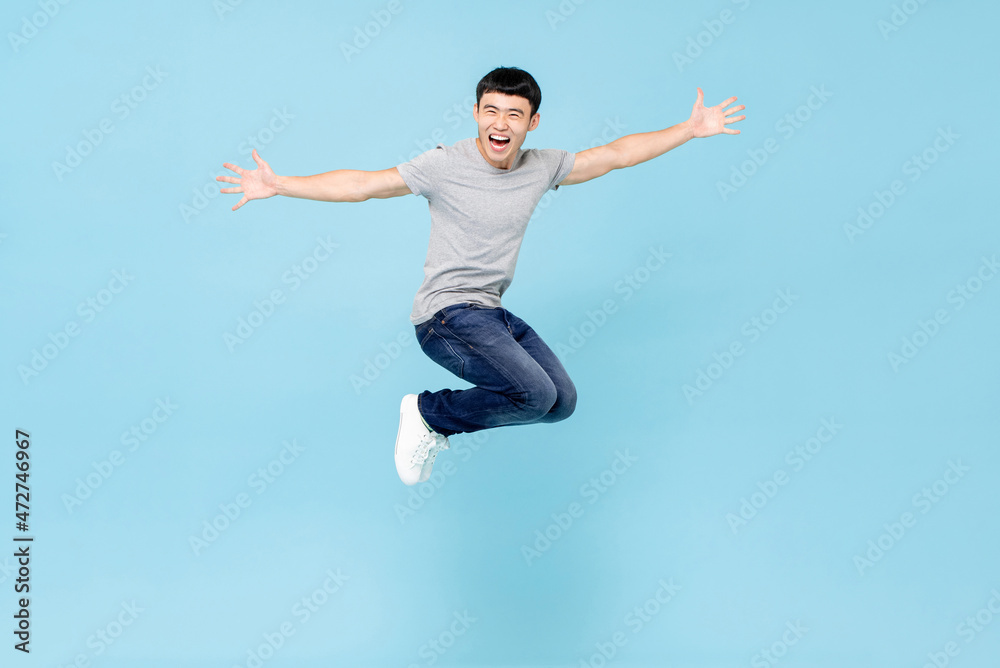 Jumping ecstatic young Asian man with arms stretching in mid air isolated in blue studio background