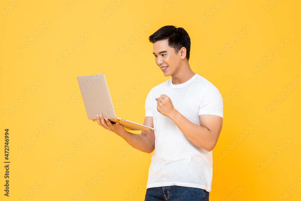 Smiling young handsome Asian man looking at laptop with winning fist gesture in isolated studio yellow background Smiling young handsome Asian man looking at laptop with winning fist gesture in isolated studio yellow background
