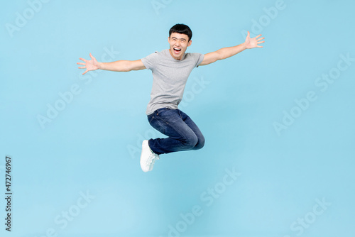 Jumping ecstatic young Asian man with arms stretching in mid air isolated in blue studio background