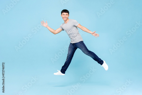 Jumping shot of ecstatic young Asian man with arms raising in mid air isolated in blue studio background