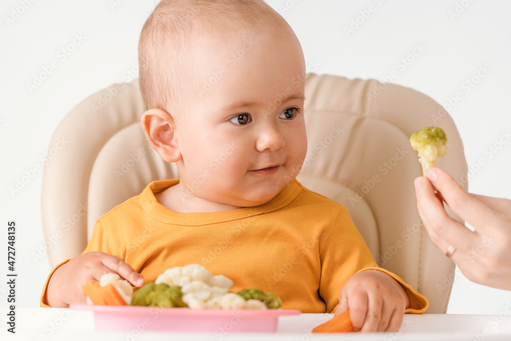 The mother's hand gives the baby sitting on a feeding chair boiled broccoli. The concept of healthy nutrition for babies The mother's hand gives the baby sitting on a feeding chair boiled broccoli. The concept of healthy nutrition for babies