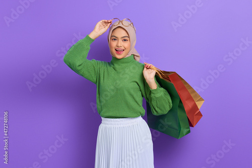 Cheerful beautiful Asian woman in green sweater and glasses holding shopping bags isolated over purple background
