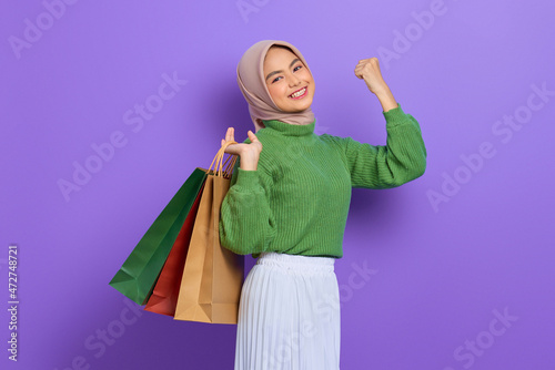 Cheerful beautiful Asian woman in green sweater holding shopping bags and celebrate luck isolated over purple background