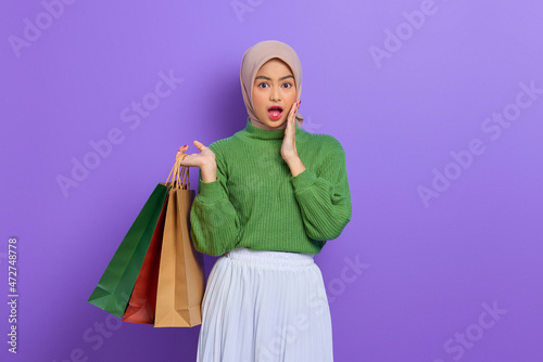 Shocked beautiful Asian woman in green sweater holding shopping bags with open mouth isolated over purple background