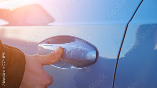 Driver in warm black jacket uses keyless entry access by putting finger on handle to open or close blue foreign car with fingerprint closeup, sunlight photo
