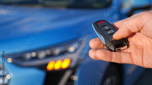 Man uses thumb to press black button on remote control key removing alarm signalling from bright blue car and headlights flash closeup © lenblr