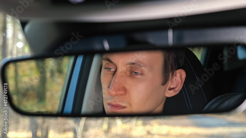 Rearview mirror reflection of dark-haired man sitting at steering wheel upset because of new car breakdown standing on forest roadside in autumn © lenblr