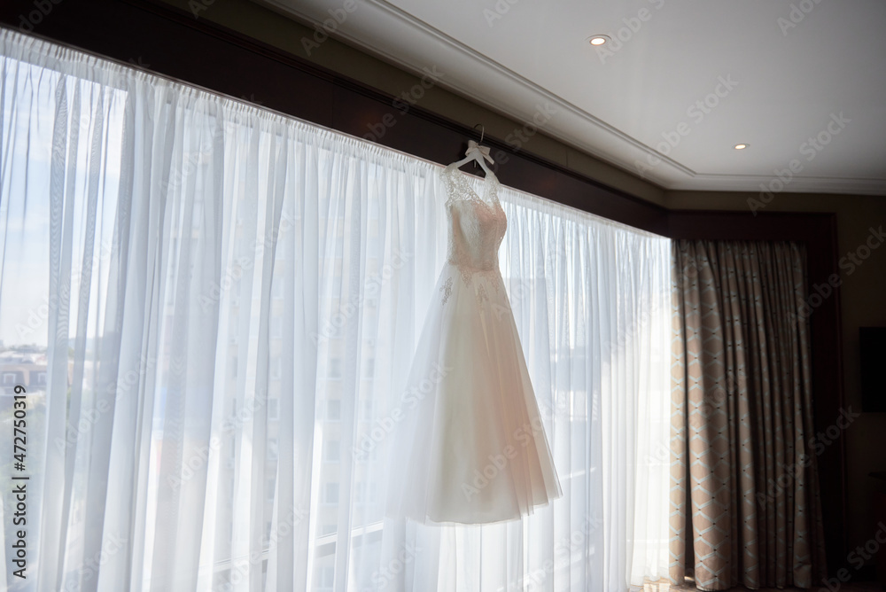Beautiful beige embroidered wedding dress hanging on hanger against window in the room, copy space, close up. Bridal morning preparations Beautiful beige embroidered wedding dress hanging on hanger against window in the room, copy space, close up. Bridal morning preparations