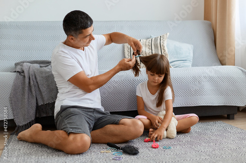 Indoor shot of young adult attractive man wearing white t shirt and jeans sitting on floor near sofa and making hairstyle for his cute daughter, preparing for going to school.